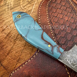 May include: A blue and brown marbled handle knife with a Damascus steel blade. The knife is resting on a brown leather sheath with decorative stitching.