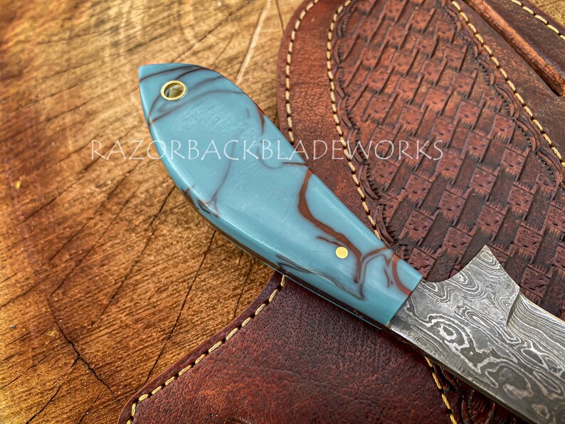 May include: A blue and brown marbled handle knife with a Damascus steel blade. The knife is resting on a brown leather sheath with decorative stitching.