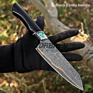 May include: A knife with a black ebony handle and a patterned Damascus steel blade featuring a copper-colored accent line. The bolster is decorated with a turquoise and silver-toned inlay. The text Black Ebony Handle appears at the top right.