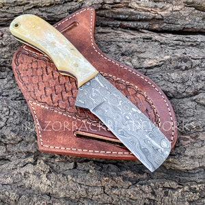 May include: A Damascus steel utility knife with a white bone handle and a leather sheath. The knife is resting in the sheath on a wooden surface.