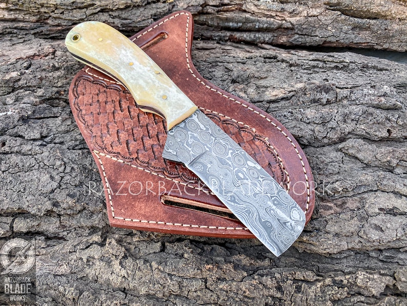 damascus cowboy bull cutter knife with a pancake leather sheath and bone handle, knife handle is assembled with brass pins and also has a lanyard hole at the bottom. the damascus pattern on the blade is raindrop style