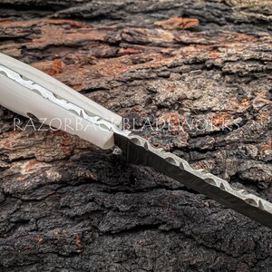 Peut inclure: Un couteau &agrave; manche blanc avec un motif d&eacute;coratif sur le manche et une lame noire &agrave; bord ondul&eacute;. Le couteau repose sur un morceau d'&eacute;corce d'arbre.