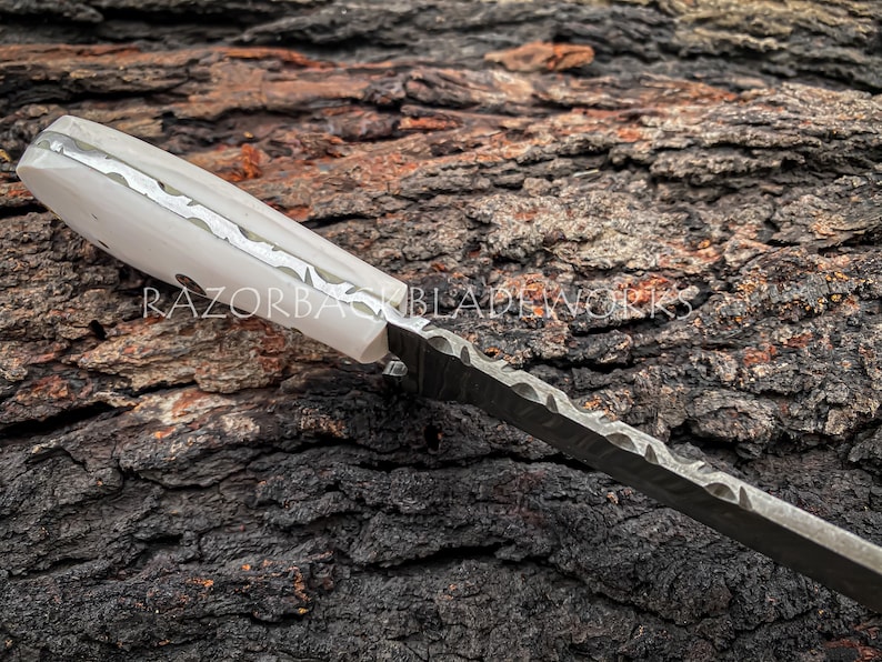 Peut inclure: Un couteau &agrave; manche blanc avec un motif d&eacute;coratif sur le manche et une lame noire &agrave; bord ondul&eacute;. Le couteau repose sur un morceau d'&eacute;corce d'arbre.
