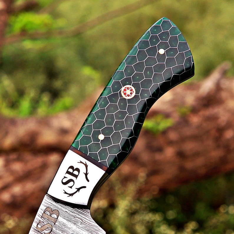 May include: A knife handle featuring a dark green resin inlay with a black honeycomb pattern. The handle includes a central decorative pin with a red floral design and two smaller white pins. A metal bolster displays the letters SB and antler icons.