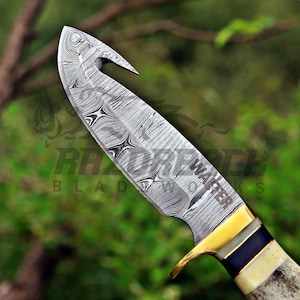 May include: A fixed-blade knife featuring a Damascus steel blade with a gut hook and the name WALTER engraved on the side. The handle includes a brass bolster, a dark blue spacer, and a textured, light-colored bone or antler grip section.