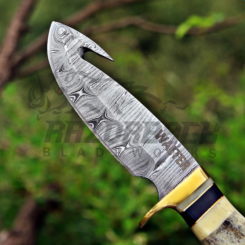 May include: A fixed-blade knife featuring a Damascus steel blade with a gut hook and the name WALTER engraved on the side. The handle includes a brass bolster, a dark blue spacer, and a textured, light-colored bone or antler grip section.