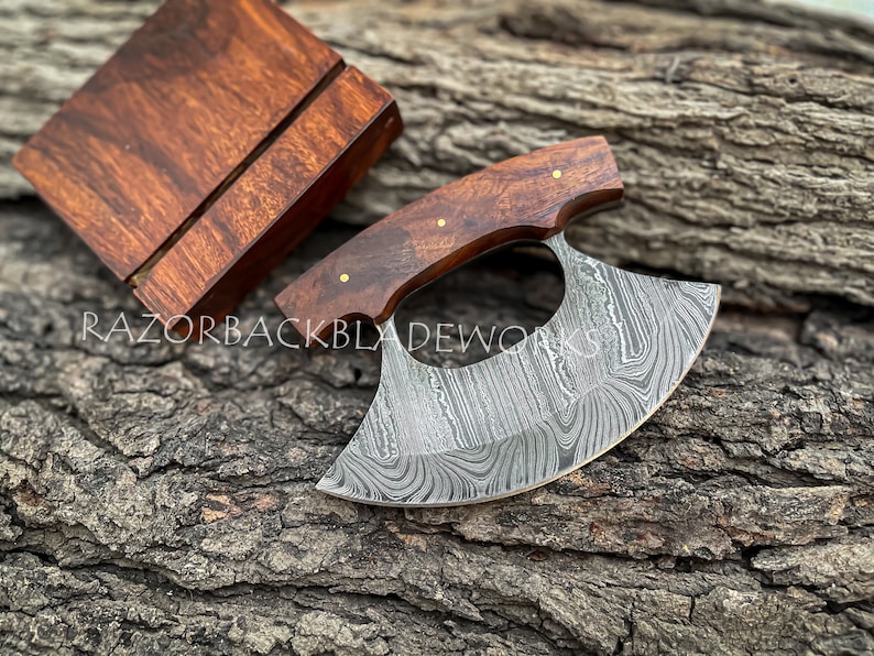 May include: A handcrafted Damascus steel ulu with a dark brown wood handle and a wooden stand to keep the ulu knife. The blade features a striking, swirling pattern. Brass pins accent the handle.  A matching wood display block is included. RazorbackBladeworks is subtly watermarked on the image. The ulu rests on a rustic, grey-brown tree trunk background.