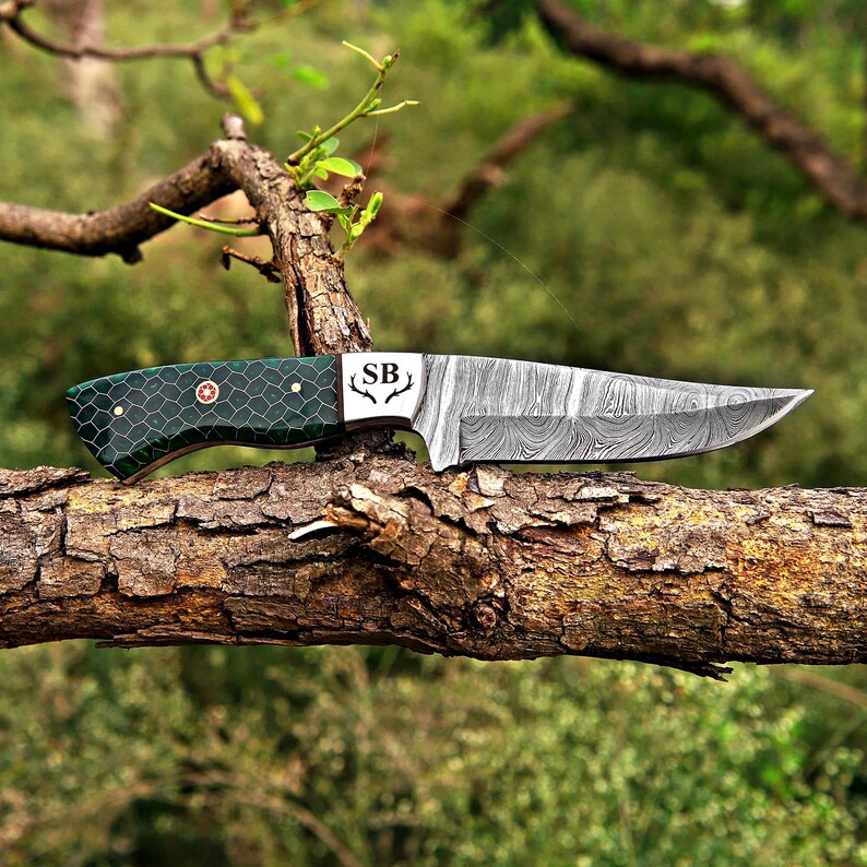 May include: A fixed-blade knife with a Damascus steel blade featuring a swirling pattern and the letters SB with an antler icon. The handle is dark green with a black honeycomb pattern, secured by three metal pins, including a central floral pin.