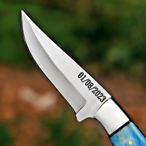 May include: A fixed-blade knife with a polished silver-colored steel blade featuring the engraved date 01/08/2023. The knife has a distinct bolster and a handle with a blue and white marbled pattern, accented by a small yellow circular inlay.