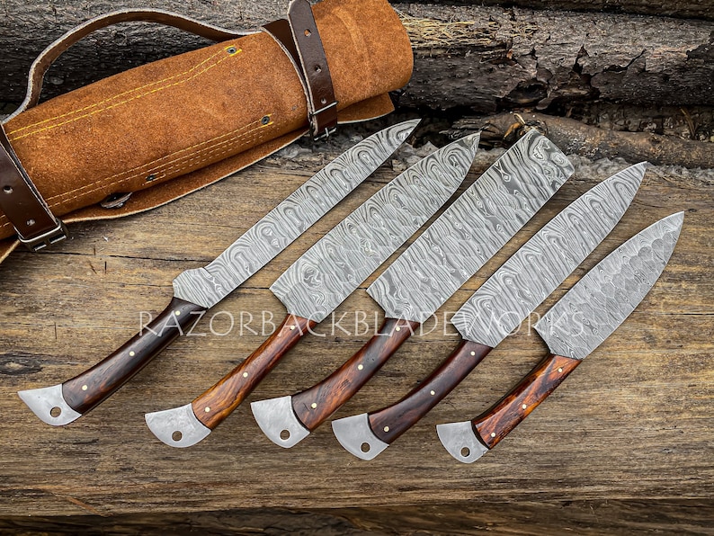 May include: Set of six Damascus steel kitchen knives with wooden handles and a leather roll for storage. The knives have a unique patterned blade and are arranged on a wooden surface.