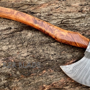 May include: A hand-forged axe with a Damascus steel blade and a wooden handle. The axe is resting on a piece of wood with the text "RZR BLADE" visible.