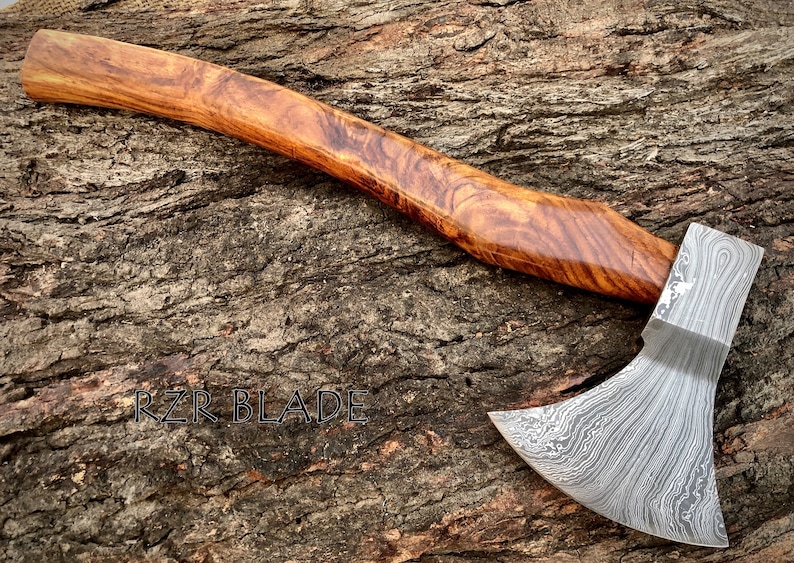 May include: A hand-forged axe with a Damascus steel blade and a wooden handle. The axe is resting on a piece of wood with the text "RZR BLADE" visible.
