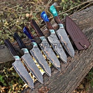 May include: Five kitchen knives with patterned Damascus steel blades and dark wood handles featuring colorful resin accents in blue, green, and teal. Each knife has a distinct handle color and a matching brown leather sheath is placed to the side.