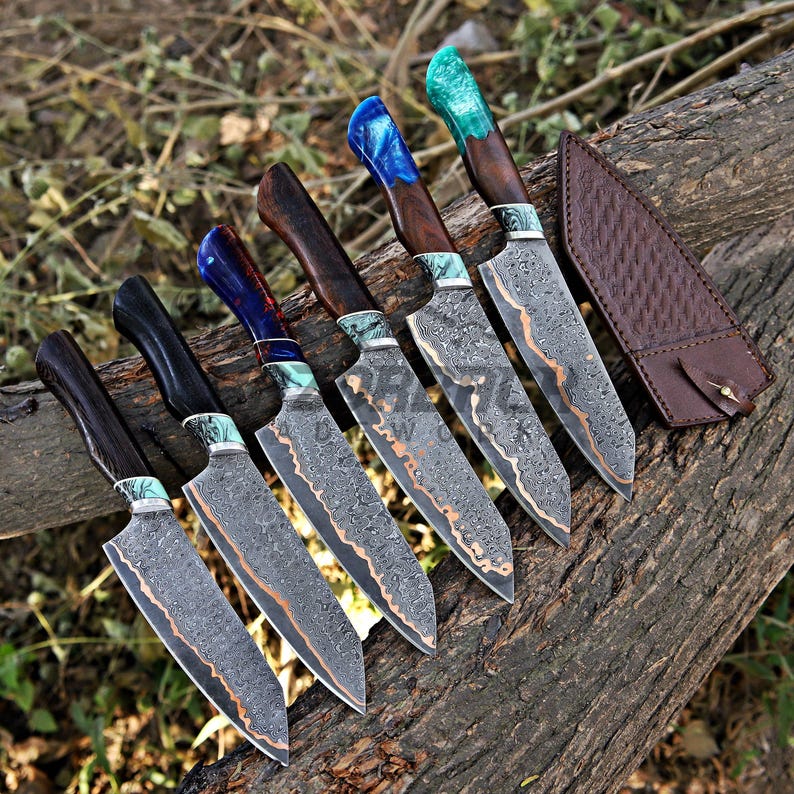 May include: Five kitchen knives with patterned Damascus steel blades and dark wood handles featuring colorful resin accents in blue, green, and teal. Each knife has a distinct handle color and a matching brown leather sheath is placed to the side.