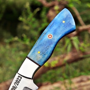May include: A knife handle featuring a vibrant blue, mottled texture with a central decorative mosaic pin surrounded by two smaller brass pins. The handle is attached to a metallic bolster with the visible text 08/2023 engraved on the blade.