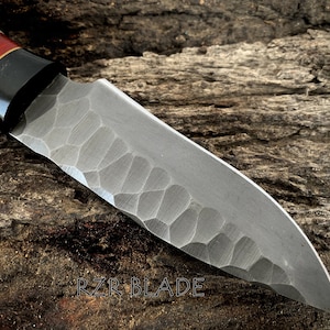 May include: Close-up of a handcrafted knife with a textured blade and a handle featuring a combination of white, red, and black materials. The blade has a unique pattern. The text "RZR BLADE" is visible.