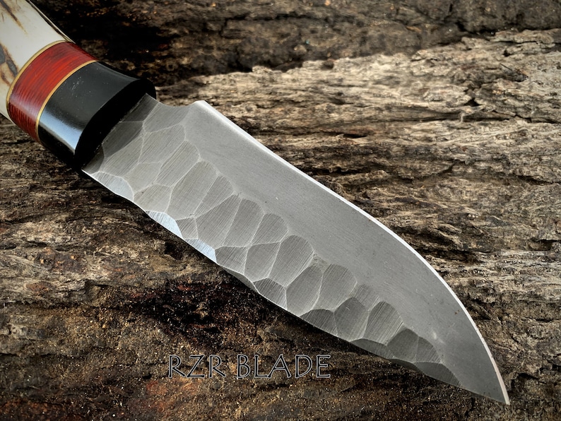 May include: Close-up of a handcrafted knife with a textured blade and a handle featuring a combination of white, red, and black materials. The blade has a unique pattern. The text "RZR BLADE" is visible.