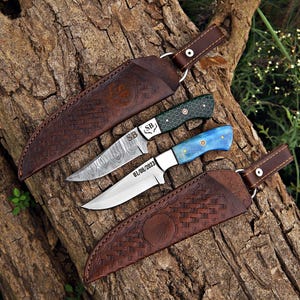 Two handcrafted fixed-blade knives on a rustic log, featuring Damascus and polished steel blades, colorful resin handles, and decorative mosaic pin details.