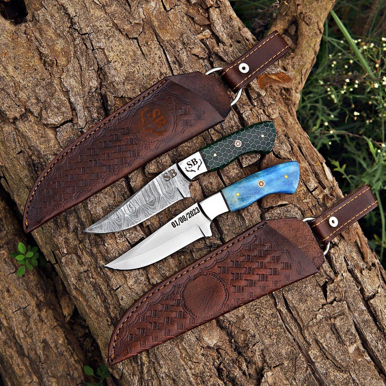 Two handcrafted fixed-blade knives on a rustic log, featuring Damascus and polished steel blades, colorful resin handles, and decorative mosaic pin details.