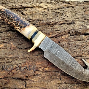 May include: A Gut hook hunting knife with a brown and black stag handle and a Damascus steel blade. The blade has a hook on the end. The knife is lying on a piece of wood with the text 'RZR BLADE' visible.