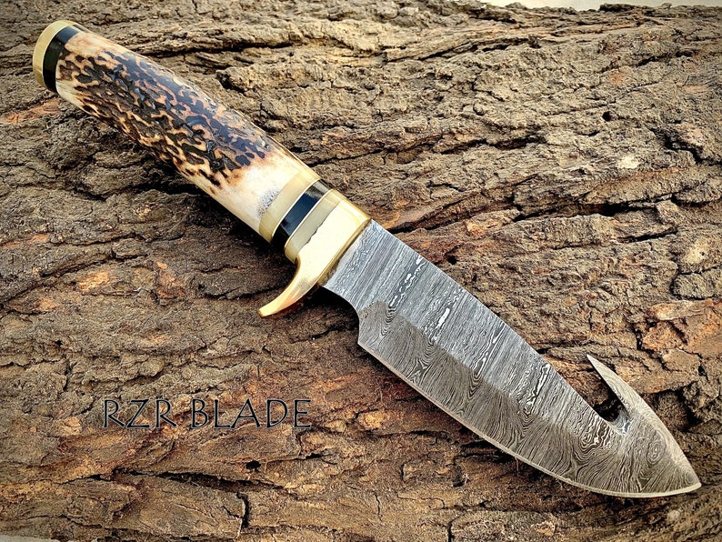 May include: A Gut hook hunting knife with a brown and black stag handle and a Damascus steel blade. The blade has a hook on the end. The knife is lying on a piece of wood with the text 'RZR BLADE' visible.