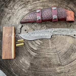 Peut inclure: Gros plan sur une lame de couteau faite &agrave; la main avec une finition &agrave; motifs. La lame repose sur une surface en bois &agrave; c&ocirc;t&eacute; d'un fourreau en cuir et d'un petit bloc de bois. Le texte "RAZORBACKBLADEWORKS" est visible sur la lame.