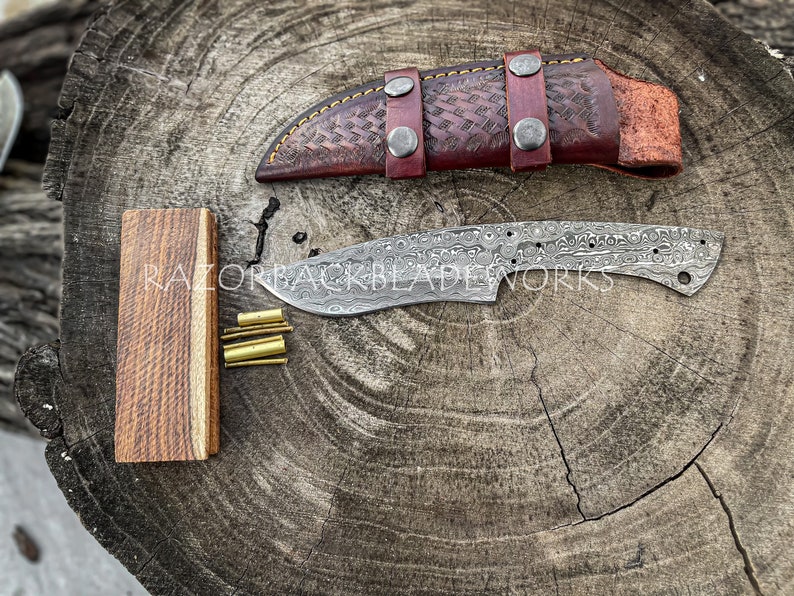 Peut inclure: Gros plan sur une lame de couteau faite &agrave; la main avec une finition &agrave; motifs. La lame repose sur une surface en bois &agrave; c&ocirc;t&eacute; d'un fourreau en cuir et d'un petit bloc de bois. Le texte "RAZORBACKBLADEWORKS" est visible sur la lame.