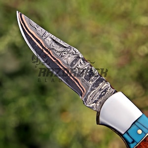 Op de afbeelding: Een zakmes met een patroon van Damascusstaal, voorzien van donkergrijze, zilveren en koperkleurige lagen. Het handvat bevat een gepolijste zilveren bolster, een fel turquoise inleg en een houten gedeelte met een messing pin.