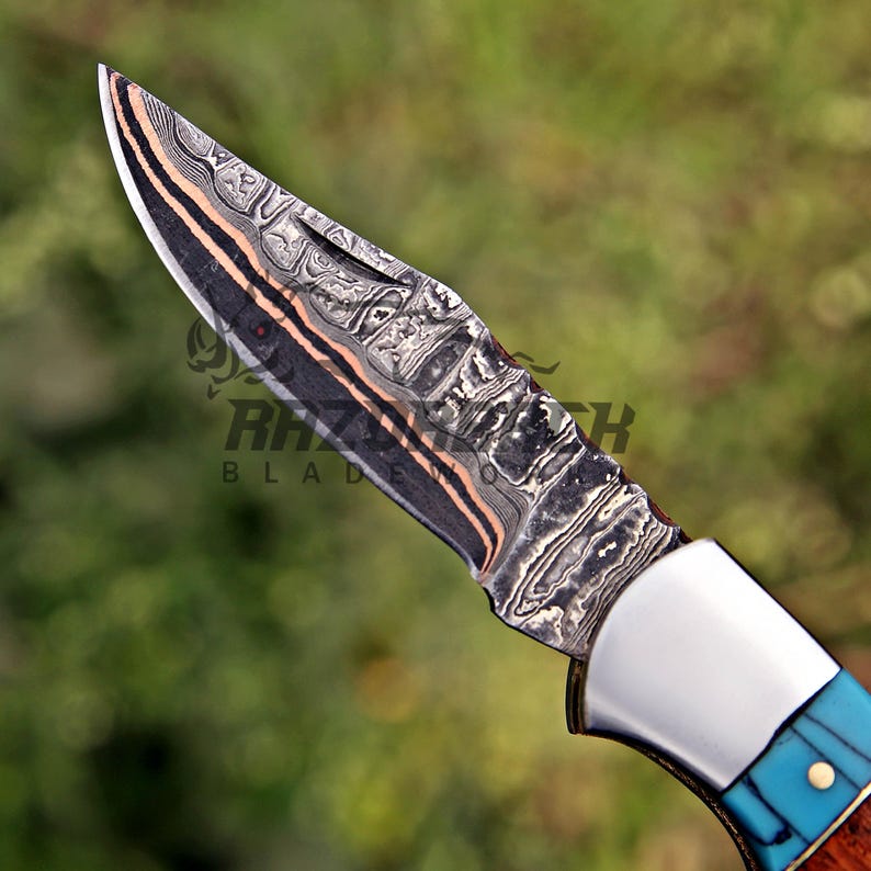 Op de afbeelding: Een zakmes met een patroon van Damascusstaal, voorzien van donkergrijze, zilveren en koperkleurige lagen. Het handvat bevat een gepolijste zilveren bolster, een fel turquoise inleg en een houten gedeelte met een messing pin.