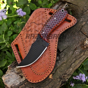 Puede incluir: Un cuchillo de caza negro y marr&oacute;n con una hoja texturizada y un mango con patr&oacute;n de panal. El cuchillo est&aacute; en una funda de cuero marr&oacute;n con costuras decorativas.
