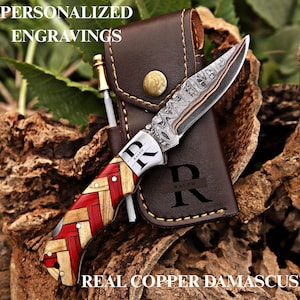 May include: A folding knife with a patterned copper Damascus steel blade and a handle featuring a woven red and light wood design. The bolster is engraved with the name RALPH. It includes a brown leather sheath with the letter R and RALPH.