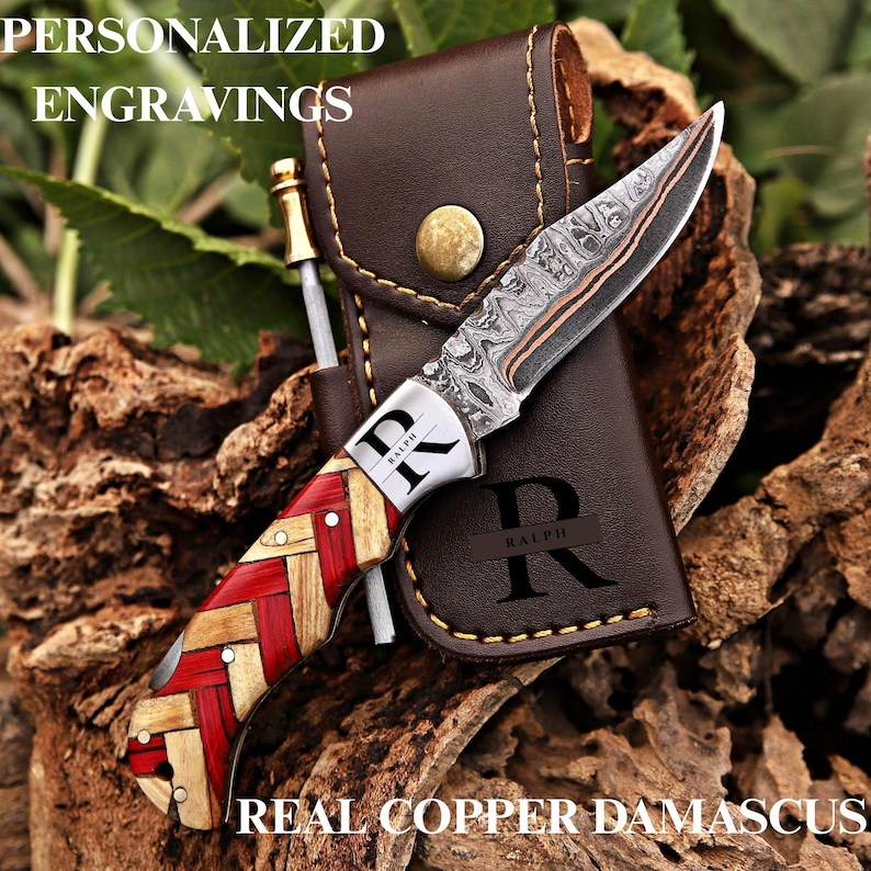 May include: A folding knife with a patterned copper Damascus steel blade and a handle featuring a woven red and light wood design. The bolster is engraved with the name RALPH. It includes a brown leather sheath with the letter R and RALPH.