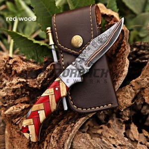 May include: A folding knife with a Damascus steel blade and a handle featuring a woven pattern of red and natural-colored wood. The knife is accompanied by a brown leather sheath with a brass snap closure and a small metal rod accessory.