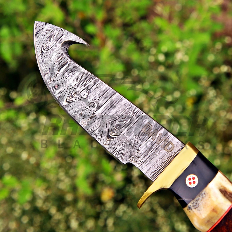 May include: A fixed-blade knife featuring a Damascus steel blade with a gut hook and a distinct swirling pattern. The handle includes a brass bolster, a black section with a circular inlay, and a textured, light-colored material at the base.