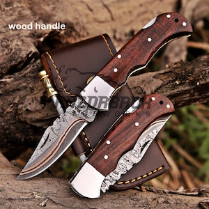 May include: Two folding pocket knives with dark brown wood handles and silver bolsters. The blades feature a distinct wavy Damascus steel pattern with a copper-colored line. One knife is open, and one is closed, resting on a brown leather sheath.