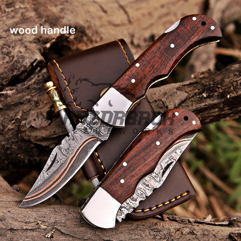 May include: Two folding pocket knives with dark brown wood handles and silver bolsters. The blades feature a distinct wavy Damascus steel pattern with a copper-colored line. One knife is open, and one is closed, resting on a brown leather sheath.