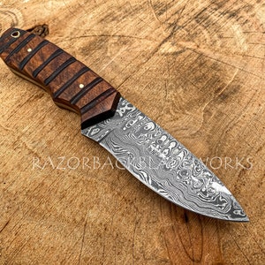 May include: A fixed-blade knife featuring a patterned Damascus steel blade and a dark brown wooden handle with horizontal grooves and brass pins. The text RAZORBACK BLADE WORKS is visible on the blade, and a logo is in the bottom left corner.