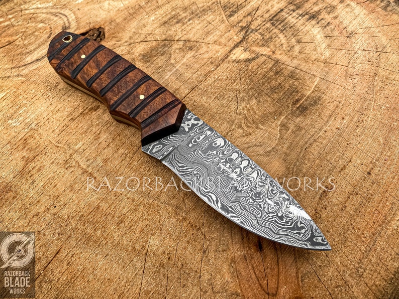 May include: A fixed-blade knife featuring a patterned Damascus steel blade and a dark brown wooden handle with horizontal grooves and brass pins. The text RAZORBACK BLADE WORKS is visible on the blade, and a logo is in the bottom left corner.