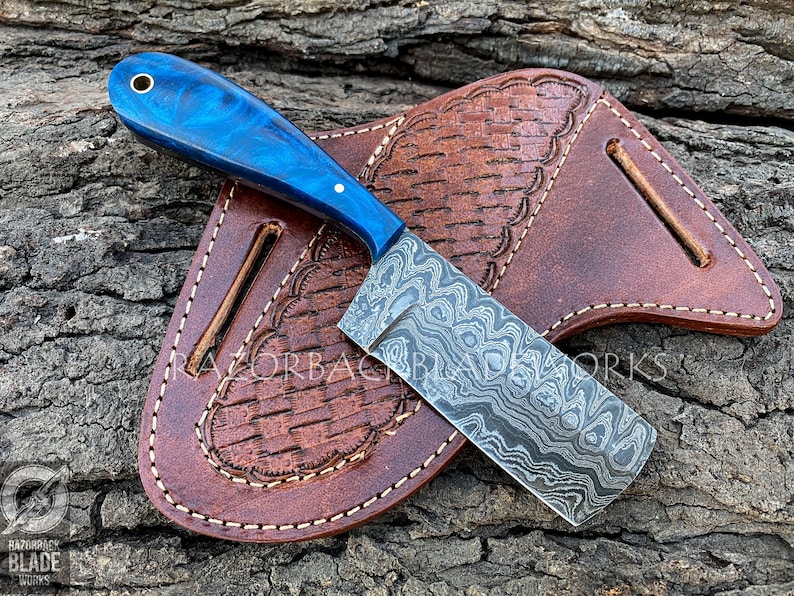 Puede incluir: Un cuchillo con patr&oacute;n azul y plateado con mango azul y funda de cuero. El cuchillo est&aacute; sobre una superficie de madera.