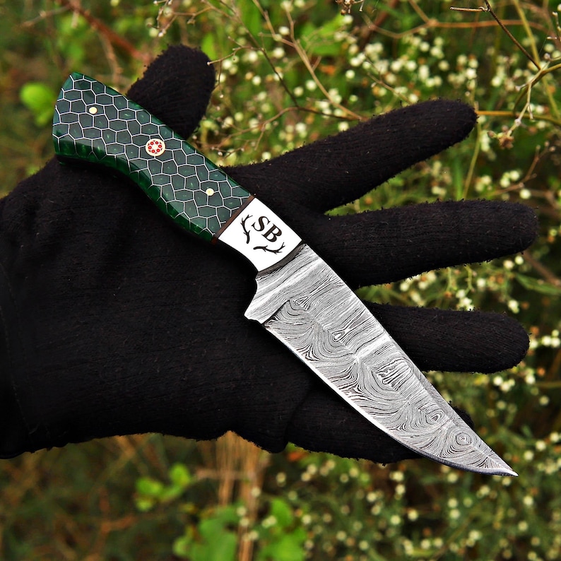 May include: A fixed-blade knife featuring a Damascus steel blade with a swirling pattern and a green handle with a black honeycomb design. The bolster is engraved with the letters SB flanked by two antler icons and secured with brass pins.