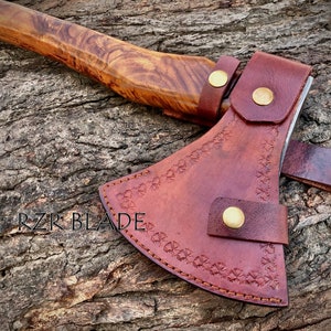 May include: A brown leather axe sheath with decorative stitching and brass snaps. The sheath is on a wooden surface. The text "RZR BLADE" is visible on the surface.