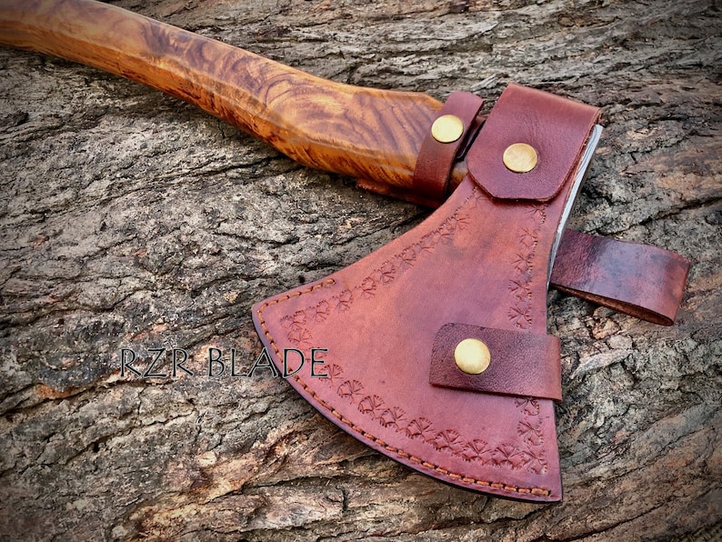May include: A brown leather axe sheath with decorative stitching and brass snaps. The sheath is on a wooden surface. The text "RZR BLADE" is visible on the surface.