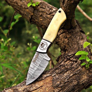 May include: A fixed-blade knife with a Damascus steel blade featuring a wavy pattern and the word Monix etched on the surface. The handle is made of a smooth, cream-colored material with two brass pins and a lanyard hole. A circular logo with the text Monix is visible on the bolster.