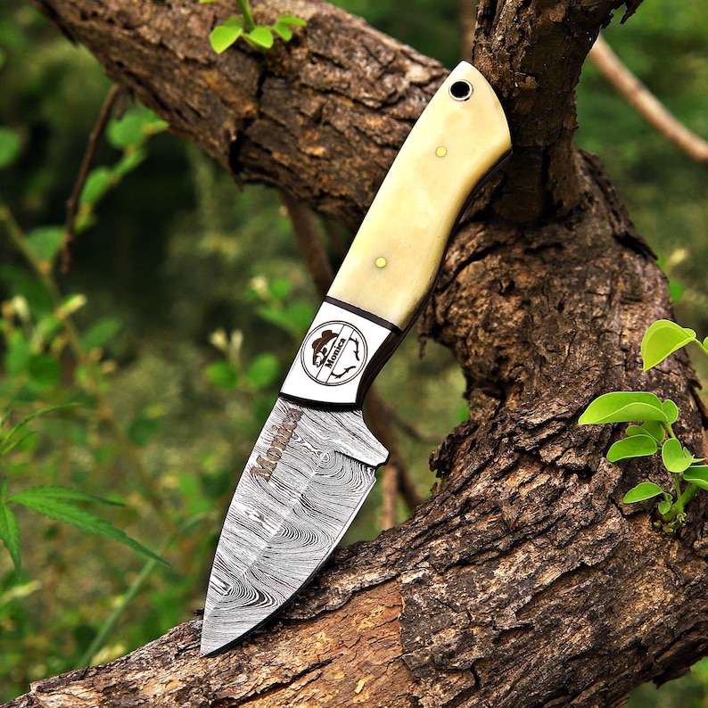 May include: A fixed-blade knife with a Damascus steel blade featuring a wavy pattern and the word Monix etched on the surface. The handle is made of a smooth, cream-colored material with two brass pins and a lanyard hole. A circular logo with the text Monix is visible on the bolster.