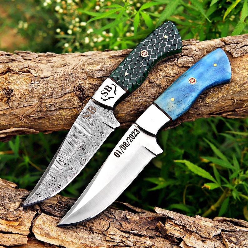 May include: Two knives with fixed blades. The top knife features a dark green handle with a honeycomb pattern and a Damascus steel blade engraved with SB. The bottom knife has a blue handle and a smooth steel blade engraved with 01/08/2023.