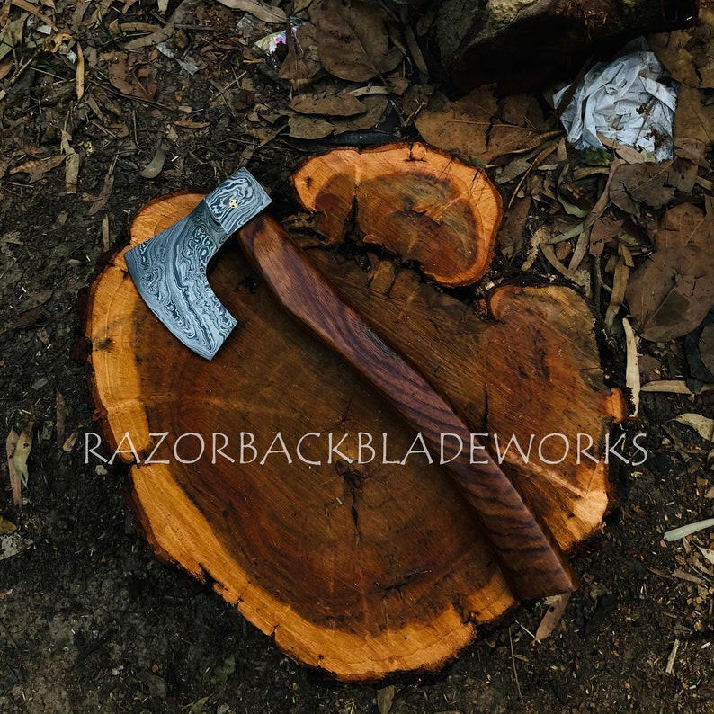 May include: A hand-forged axe with a Damascus steel blade and a wooden handle. The axe is resting on a wooden stump. The text "RAZORBACKBLADEWORKS" is visible on the stump.