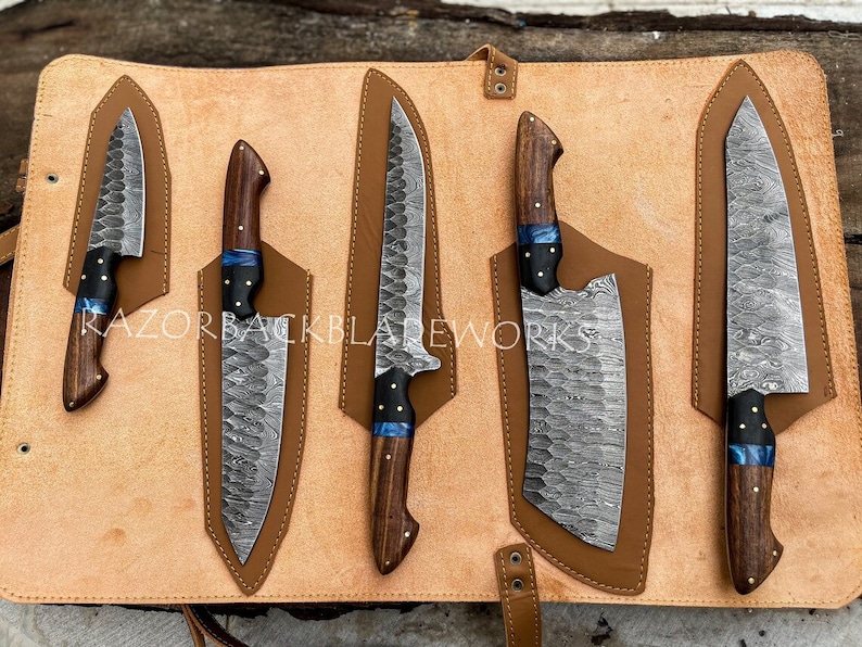 May include: Set of five kitchen knives with wooden handles and a blue accent. The knives are arranged in a leather roll with individual slots for each knife. The knives have a Damascus steel pattern.