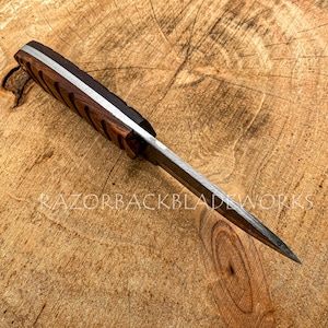 May include: A fixed-blade knife with a dark brown wooden handle featuring diagonal grooves and a silver-colored metal spine. The blade is pointed and metallic. The text RAZORBACKBLADEWORKS is visible on the surface, along with a circular logo.