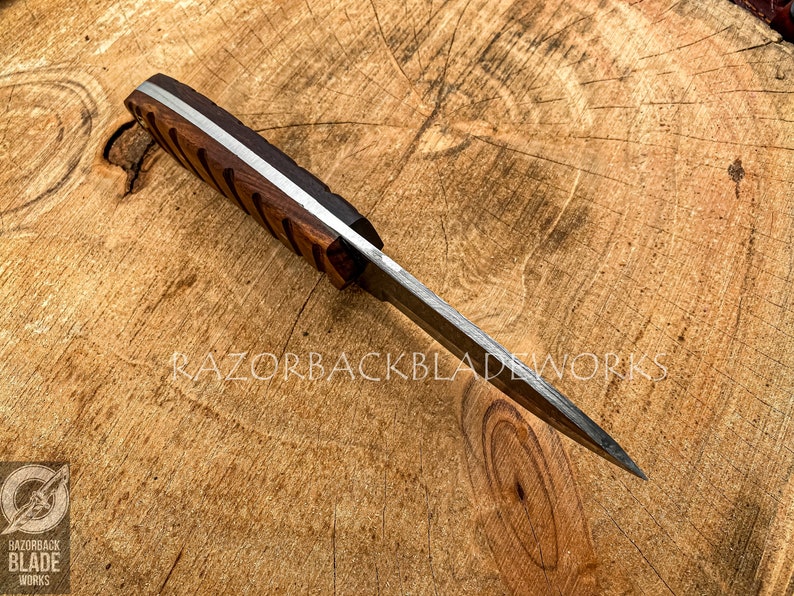 May include: A fixed-blade knife with a dark brown wooden handle featuring diagonal grooves and a silver-colored metal spine. The blade is pointed and metallic. The text RAZORBACKBLADEWORKS is visible on the surface, along with a circular logo.