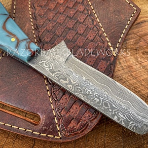 May include: A straight razor with a blue and brown marbled handle and a damascus steel blade. The razor is resting on a brown leather sheath with decorative stitching.